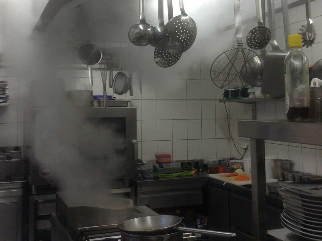 Gewerbliche Küche mit Dampfschwaden unter der Abzugshaube, Kochlöffel und Kellen hängen an der Haube